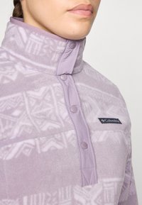 Jasnofioletowy polarowy półgolf z wysokim kołnierzem, zapięciem na napy oraz geometrzycznym wzorem. Posiada małe logo na obszarze klatki piersiowej.