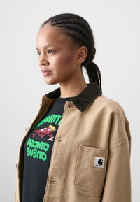 Tan cotton jacket with a dark corduroy collar, featuring a patch pocket and logo. Black T-shirt beneath has colorful graphic text and cartoon.