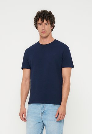 Hombre con cabello rizado y oscuro, vistiendo una camiseta lisa azul marino de manga corta y vaqueros azul claro, de pie contra un fondo blanco.