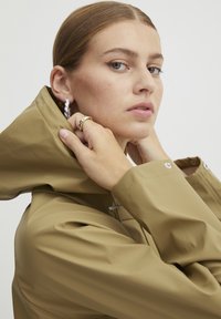 Kahaki regnkappa med hög krage, slät vattentålig textur och knappdetaljer. Modellen bär silverfärgade hoopörhängen och ringar.