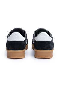 Schwarze Schuhe aus Wildleder und Leder mit weißen Akzenten, ausgestattet mit einer Gummisohle. Die Ferse zeigt ein geprägtes Logo. Rückansicht, minimalistische Details.