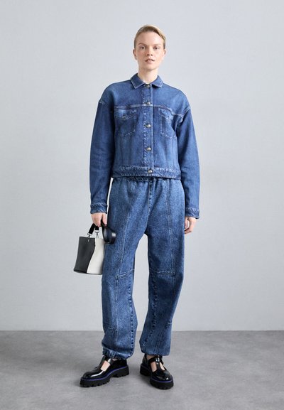 Jachetă din denim cu guler și închidere buton, asociată cu pantaloni din denim largi și conici. Modelul ține o mică geantă de mână alb-negru.