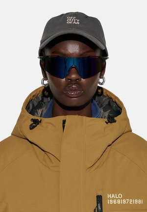 Person trägt dunkle Wraparound-Sonnenbrille, silberne Creolen, dunkelgraue Kappe mit der Aufschrift "OFF DUTY GEAR" und beige Kapuzenjacke mit Reißverschlusstaschen.