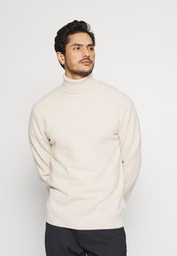 Suéter de cuello alto color beige claro, textura suave, corte relajado, con cuello y dobladillo acanalados, combinado con pantalones oscuros, de color sólido.