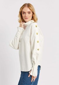 Blond naine, kes kannab valget pika varrukaga toppi, millel on kuldsed nööbid õlgadel ja sidumisvarrukad, mille alla on sobitatud sinised teksapüksid.