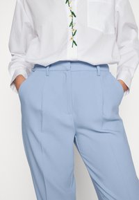 Chemise blanche boutonnée avec broderie florale verte et jaune, associée à un pantalon taille haute bleu clair, mains dans les poches.