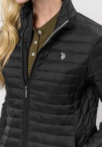 Svart vadderad jacka med dragkedja framtill, horisontellt quiltmönster och silverlogotyp på bröstet. Bärs över en olivgrön skjorta med krage.