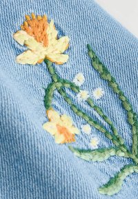 Design floral brodé mettant en avant des fleurs jaunes et orange avec des tiges vertes sur un fond en jean bleu clair. Détails de couture texturés visibles.
