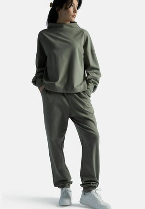 Nainen seisoo käsivarret taskuissa, yllään oliivinvihreä korkeakauluksinen collegepaita, siihen sopivat housut ja valkoiset tennarit, katsoo oikealle.