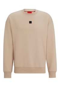 Sudadera beige con cuello redondo, puños acanalados y dobladillo. Presenta un pequeño parche de logo negro en el centro del pecho. Textura suave y lisa.
