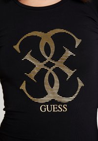 Schwarzes, figurbetontes T-Shirt mit einem großen, goldgestreiften Logo-Design, darunter in fetter Schrift das Wort "GUESS". Glatte Stofftextur.