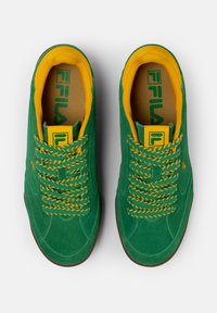 Zapatillas de ante verde con acentos amarillos. Las características incluyen cordones y logotipo amarillos, superficies texturizadas y una suela de goma marrón.