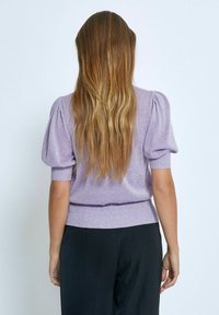 Lila puffärmad tröja med ribbad nederkant, med en glansig yta. Stylad med svarta byxor, sedd bakifrån.