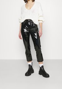 Schwarze glänzende Vinyl-Hosen mit hohem Bund und cropped Schnitt, kombiniert mit einem locker sitzenden weißen Strickpullover und klobigen schwarzen Stiefeletten.