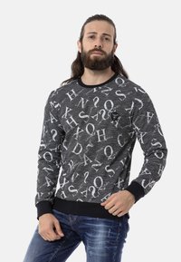 Zwarte sweatshirt met witte alfabetische print, ribbelboorden en -zoom, en een logopatch op de borst, gecombineerd met blauwe jeans.
