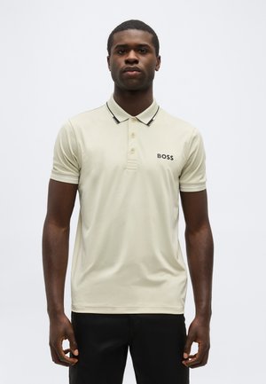 Uomo che indossa una polo BOSS beige con rifiniture nere sul colletto e pantaloni neri, in piedi davanti a uno sfondo chiaro e uniforme.