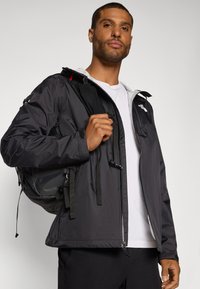 Schwarze leichte Jacke mit grauem Kapuzen, ausgestattet mit Reißverschlusstaschen und Kordelzügen. Model hält einen schwarzen Rucksack über einem weißen Shirt.