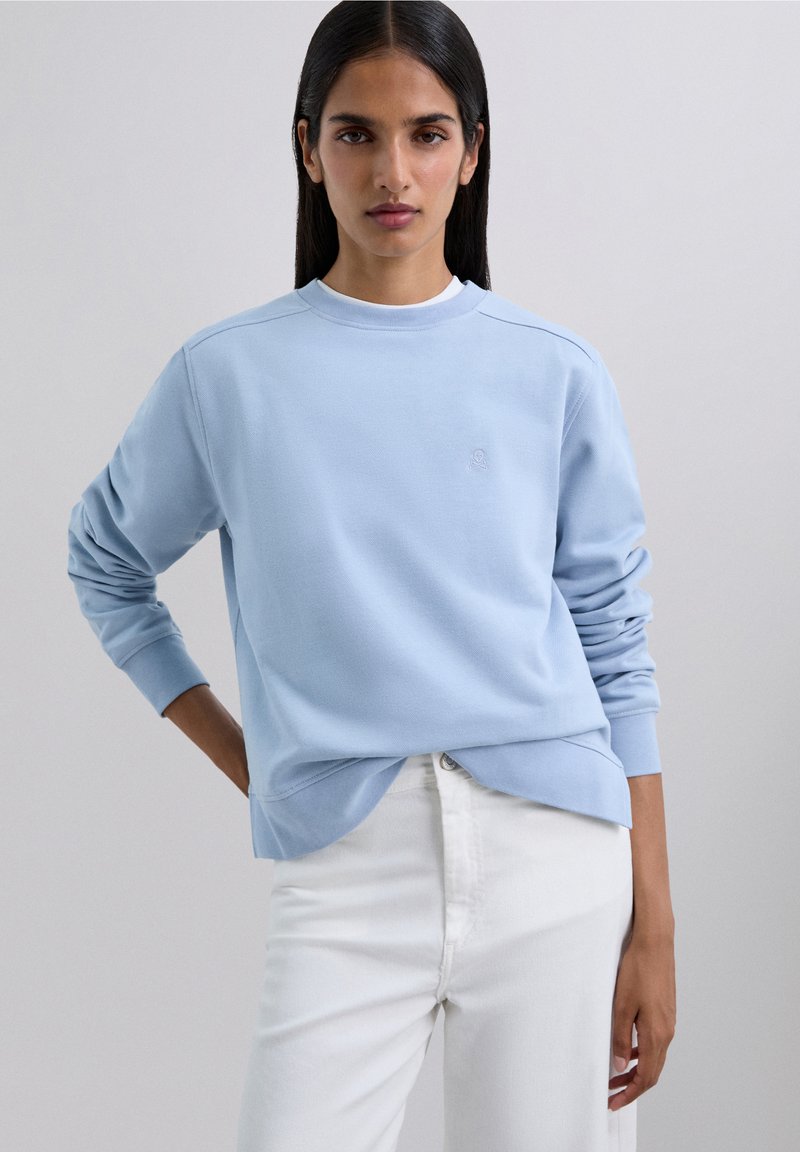 Sudadera azul claro con cuello redondo y puños acanalados. Presenta un pequeño logo bordado en el pecho. Combinada con jeans blancos.
