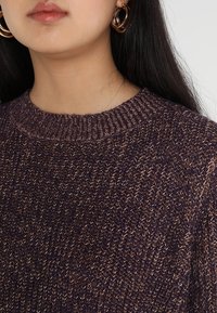 Gestrickter lila Pullover mit strukturierter Oberfläche und geripptem Ausschnitt. Modell trägt große Creolen. Nahaufnahme von Stoffdetails.