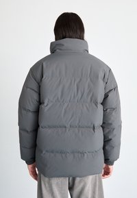 Chaqueta de plumas gris con un cuello alto, con amplias secciones acolchadas horizontales, textura suave y puños elásticos en las muñecas.