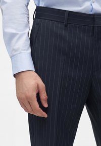 Άνδρας που φοράει σκούρο μπλε παντελόνι με ρίγες και ανοιχτό μπλε πουκάμισο με μακριά μανίκια, με εμφανές χέρι στο πλάι.