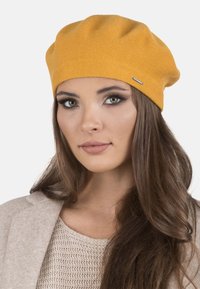 Beret de punto amarillo con una textura suave, que presenta una pequeña etiqueta de marca metálica. El diseño tiene un ajuste holgado, creando una silueta relajada.