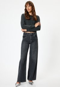 Jeans larghi grigi scuri con vita alta, chiusura con bottone e zip, e tasche laterali. Abbinati a un maglione corto e button-up scuro.