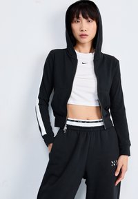 Sudadera negra con cremallera y franjas laterales blancas, combinada con un top corto blanco y pantalones de chándal negros con cintura y logo de la marca.