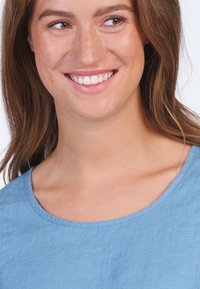 Helles blaues, kurzärmliges Oberteil mit rundem Ausschnitt. Der Stoff wirkt weich mit einer glatten Textur. Das Model lächelt und zeigt klare Haut und Zähne.