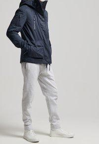 Osoba w granatowej kurtce z kapturem i kieszeniami, jasnoszarych dresach oraz białych sneakersach, stojąca na jednolitym jasnym tle.