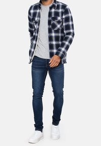 Chemise en flanelle à carreaux bleu marine et blanc avec deux poches poitrine, portée sur un t-shirt gris, associée à un jean skinny foncé et des baskets blanches.