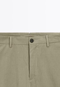 Närbild av midjan på beige byxor med bältesögglor, knappstängning och sömnad på gylfen mot en vit bakgrund.