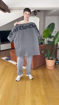 Homem a standing numa sala de estar segurando uma grande t-shirt cinza com texto gótico preto, vestindo tênis brancos e calças cinzas.