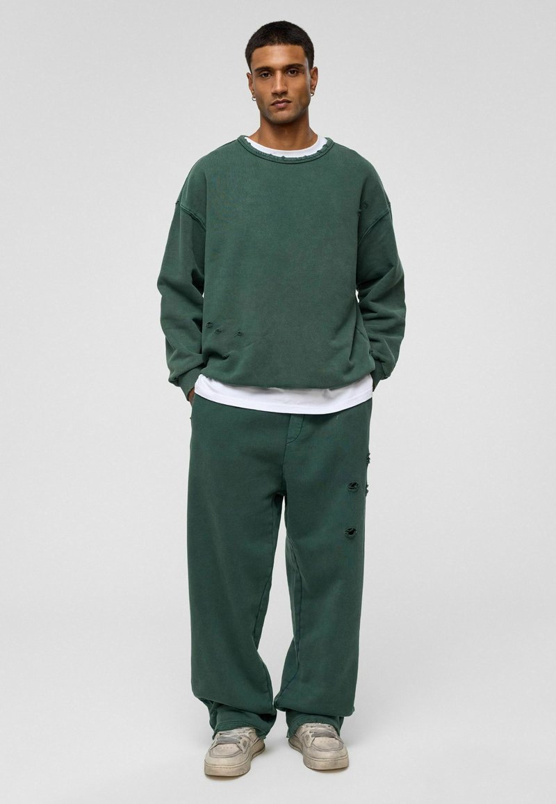 Groene katoenen sweatshirt met versleten details, gedragen over een wit T-shirt, gecombineerd met bijpassende groene broek. Model staat voor een neutrale achtergrond.