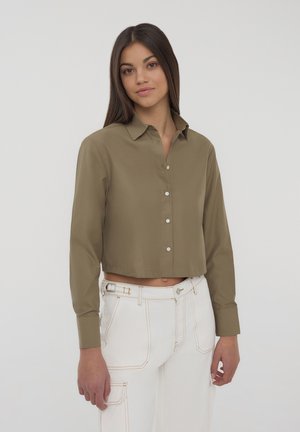 Camicia verde oliva tagliata, con colletto, maniche lunghe e bottoni bianchi, abbinata a pantaloni bianchi a vita alta con cuciture arancioni.