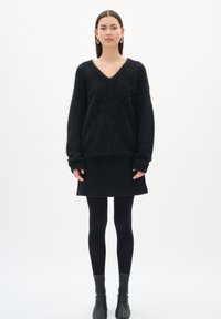 Žena oblečená v čiernom huňatom svetri s výstrihom do V, čiernej mini sukni, čiernych pančuškách a čiernych členkových topánkach, stojí oproti bielej stene.