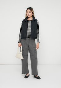 Ung kvinna som bär en svart pufferväst, svart-vit randig långärmad tröja, grå cargo-byxor, svarta flats, håller en liten beige handväska.