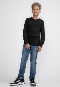 Schwarzer V-Ausschnitt-Pullover kombiniert mit hellblauen Jeans und schwarzen Sneakers mit weißen Akzenten. Das Modell steht mit entspannten Händen.