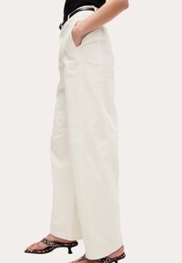 Wijdvallende witte katoenen broek met zijzakken, hoge taille en zichtbare naden. Gecombineerd met zwarte en gemêleerde sandalen met hakken en bandjes.