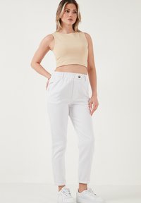 Bež cropped tank top, mille valgete kitsaste pükste jaoks, millel on elastne vöökoht ja ees nupud, kandes valgetel tossudel.