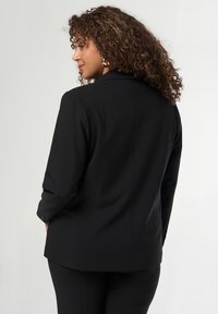 Zwarte op maat gemaakte blazer met lange mouwen, gestructureerde schouders en een gladde textuur. Beschikt over een enkele rugventilatie voor gemakkelijke beweging.