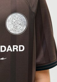 Jersey deportivo marrón con un diseño texturizado, un emblema plateado redondo en el pecho y letras blancas que dicen "STANDARD."