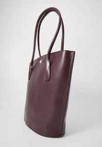 Bordeaux leren tote bag met een gladde textuur, langwerpig ontwerp en dubbele handvatten. Heeft een klein gouden logo aan de voorkant.