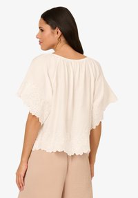 Blusa color crema con ricami sulle maniche e sull'orlo, caratterizzata da una vestibilità ampia, maniche corte a sbuffo e scollo a cuore sul retro.