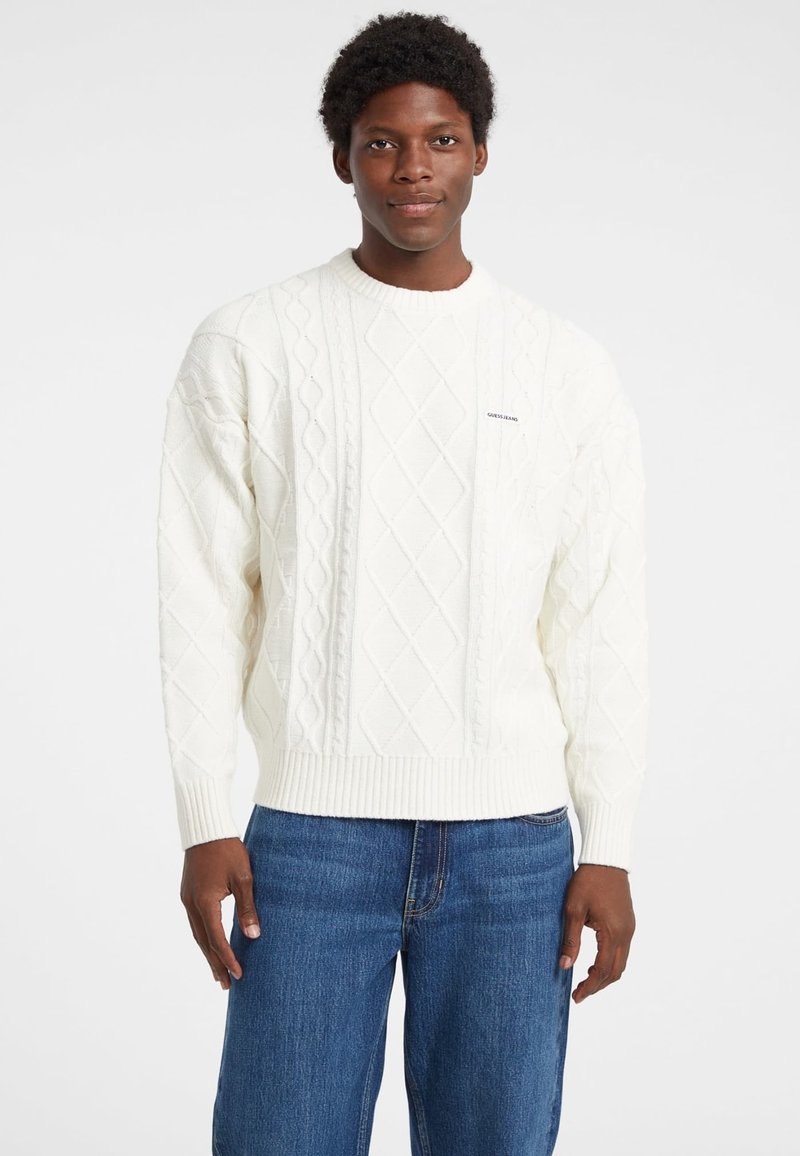 Maglione bianco a maglia con punto a coste nell'orlo e nei polsini, con motivo a rombi, indossato sopra jeans in denim blu. Logo del marchio sul petto.