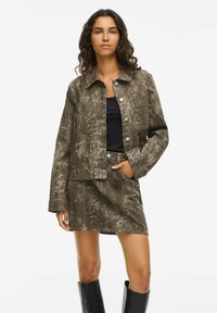 Veste imprimée serpent marron avec un col et une fermeture à boutons, assortie d'une jupe. Le mannequin porte un haut noir et des bottes à genoux.