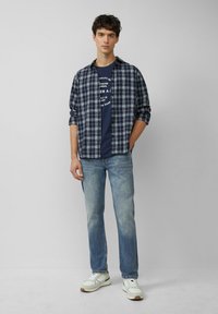 Flanellen shirt met blauw-wit plaid patroon bovenop een marineblauw t-shirt, gecombineerd met lichte spijkerbroek en witte sneakers. Eenvoudige, casual uitstraling.