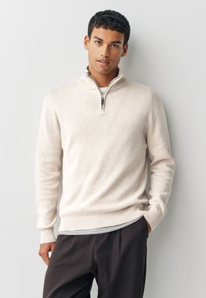 Noor mees lühikeste lokkis juustega, kandes helebeeži kvartstõmblukuga kampsunit triibulise särgi peal, seisab ühevärvilise heleda tausta ees.