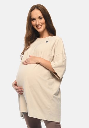Pregnant woman in loose beige dress gently holding her belly, smiling against a plain light background.