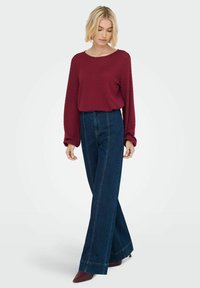 Maglione borgogna a texture con maniche lunghe, abbinato a pantaloni a vita alta in denim scuro a gamba larga, con dettagli di cucitura visibili.